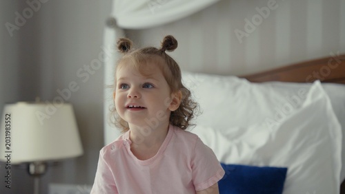 Photography Little girl with pigtails sitting on a comfortable bed, smiling as she enjoys a