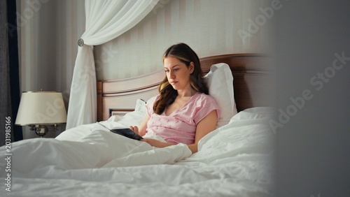 Foto Young woman in pink pajamas relaxing in a luxurious bed, reading an e reader tab