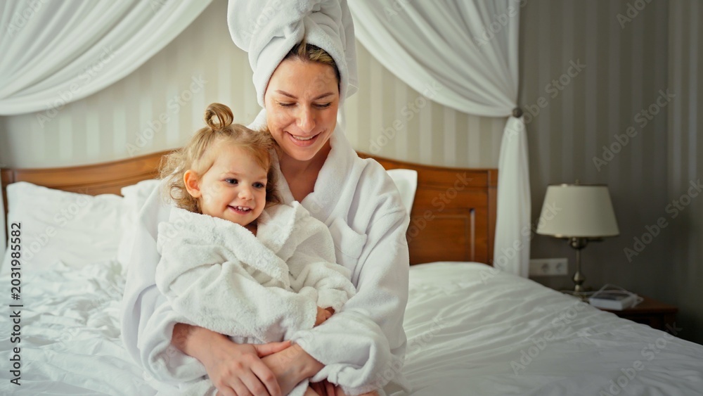 Fototapeta premium Mother and daughter wearing cozy white bathrobes and towels, smiling and embracing while sitting on a comfortable bed in a bright bedroom, starting a relaxed morning routine together