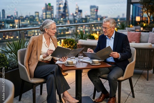 Elegant senior couple dining at rooftop restaurant with city skyline view during evening, enjoying meals and conversation.