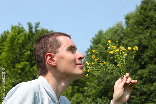 Young man profile face with small yellow flowers in hand on a green forest and sky background.