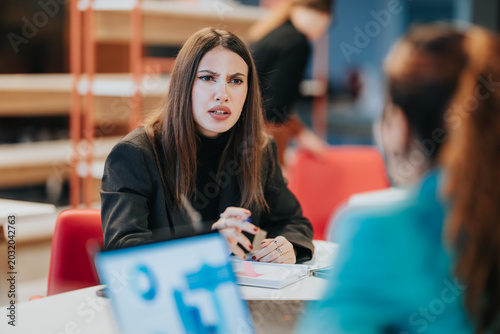 Photos A businesswoman speaks with a concerned expression while discussing documents across a table