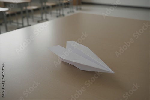 A classic dart-style paper airplane lies flat on a light brown wooden table in a quiet classroom setting.