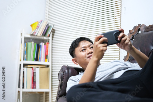 Cuadro en lienzo A cheerful Asian man is having fun playing video games on his smartphone while comfortably lying on a sofa