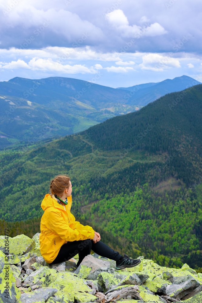 Fototapeta premium Woman hiking in mountains. Woman in yellow on the rocky mountain peak on the background of green mountain forests. Green tourism, hiking concept. Close up woman hiking. Hiking tourism advertisment. 