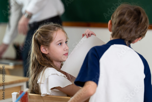 Elementary girl and boy exchange worksheet while adult male teacher moves through classroom, back to school group activity supports peer interaction, confidence, participation, learning routine.