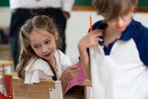 Εκτύπωση καμβά Elementary girl smiles while classmate holds pencil and worksheet near desk, back to school classroom activity builds friendship, participation, study confidence, peer learning, social skill
