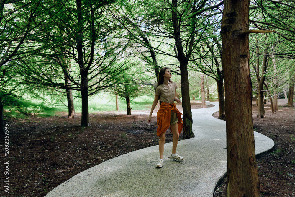 Fototapeta premium Woman walking on winding path through forest park among tall trees and nature, casual outfit with jacket tied at waist, looking up during a relaxed outdoor stroll.