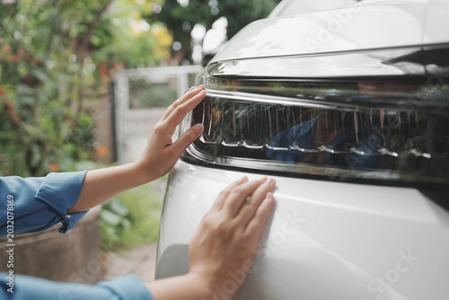 Billede på lærred Close-up of hands touching the headlight and hood of a white car outdoors, checking vehicle condition or cleaning with soft daylight and blurred garden background