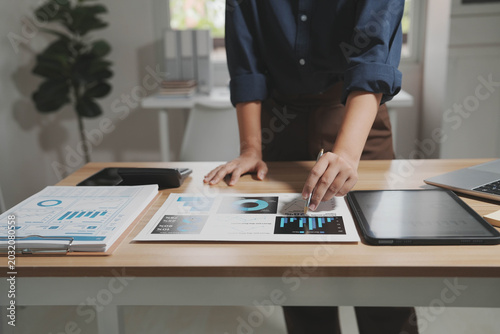 Business professional analyzing printed charts and digital dashboard at modern office desk with tablet and laptop, data and financial planning concept.