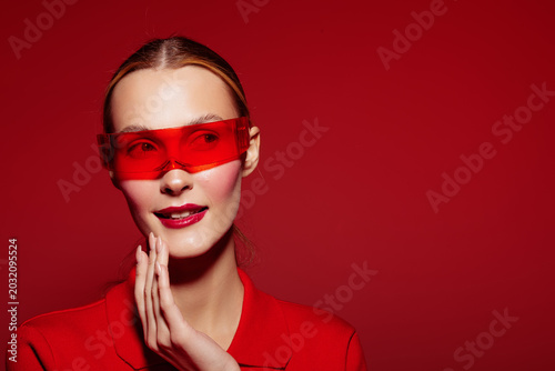 Young woman in red outfit and red sunglasses poses with hand near face on red background. Fashion portrait with vibrant colors and confident expression for style concept.