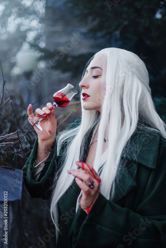 A gothic vampire with bloody hands and a mystical look drinks from a glass.