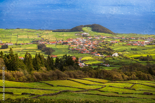 Elevated view of a small coastal village surrounded by vibrant green patchwork agricultural fields and traditional volcanic stone walls on Terceira Island in the Azores, Portugal.