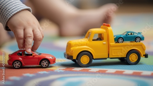A child's hand interacts with toy cars, including a towing truck transporting smaller ones.