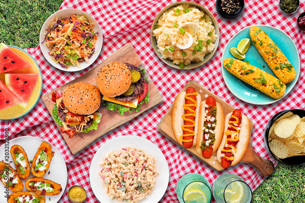 Obraz premium Outdoor summer food BBQ meal with hot dogs, hamburger and sides. Top down view on a red and white check cloth and grass background.