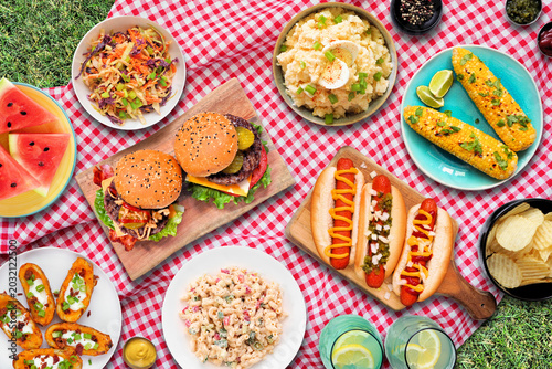 Outdoor summer food BBQ meal with hot dogs, hamburger and sides. Top down view on a red and white check cloth and grass background.