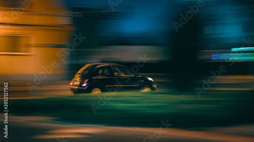 Fotografija Dark small car travels quickly on an urban street at night Buildings blurred by