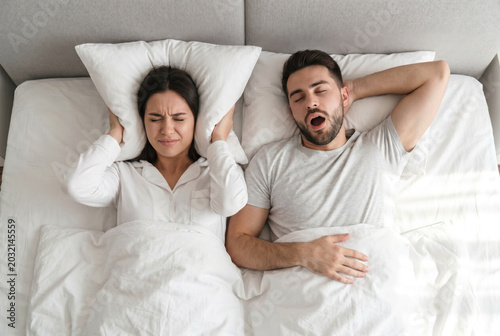 Frustrated young woman covering ears with pillow while her husband snoring loudly in bed top view