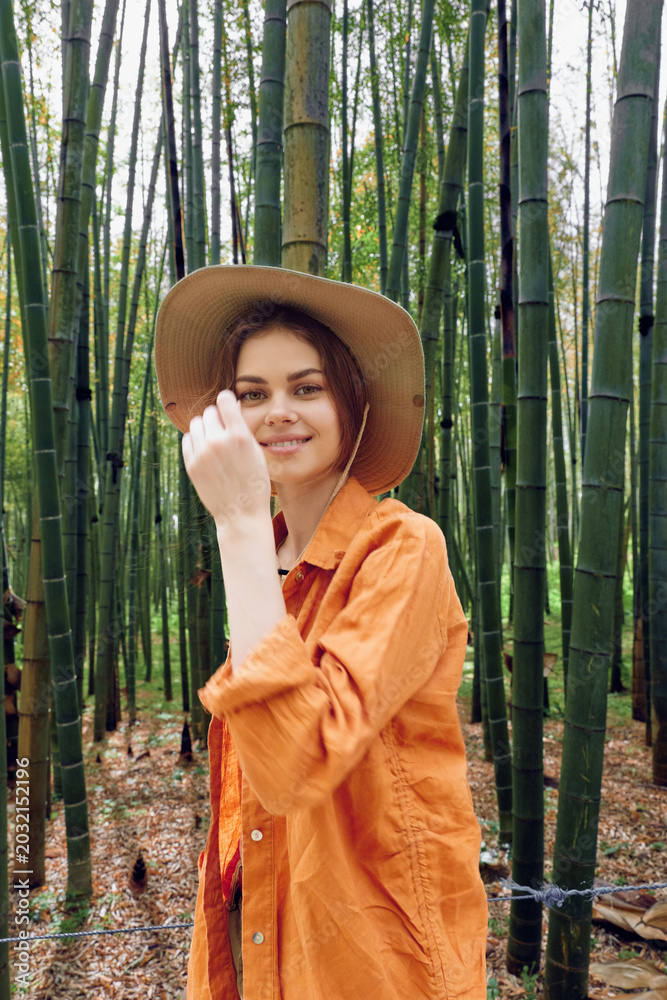 Fototapeta premium Woman in bamboo forest wearing straw hat and orange shirt, smiling in portrait style. Nature travel scene with relaxed pose among tall green stalks and serene outdoor greenery.