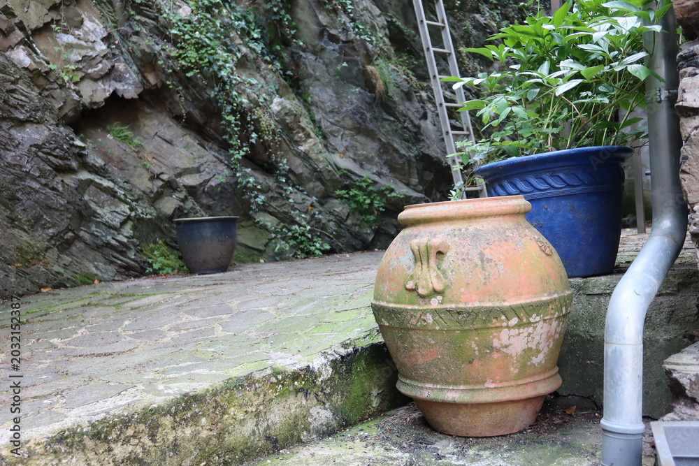 Obraz premium An old decorative clay pot stands near a stone wall in a quiet corner of Cochem, Germany. Green plants and rustic textures create a peaceful atmosphere.