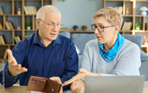 Senior couple budgeting debt pension stress wallet laptop. Couple review bills at home, showing cash shortage and rising expenses. Clear concept of financial pressure in retirement today.