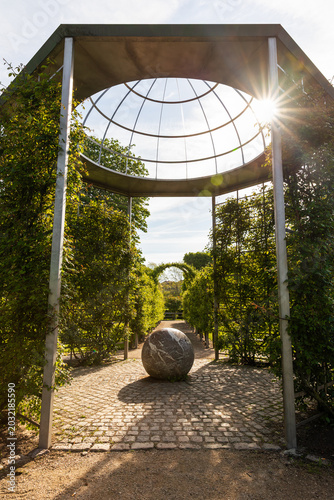 Geometric garden pavilion with a stone sphere in the King's Garden, Copenhagen. Symmetric park architecture with bright sun starburst flare in spring.