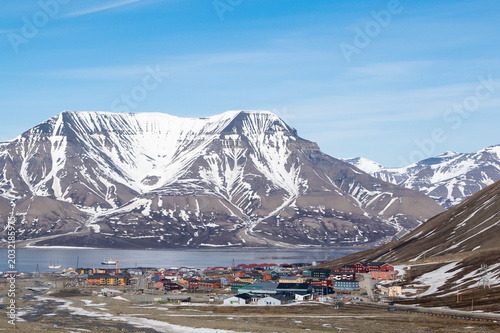 A colorful town sits nestled at the base of snow-capped mountains on the coast of Svalbard, Norway. Ships are visible in the bay, suggesting a connection to maritime activities or transport.