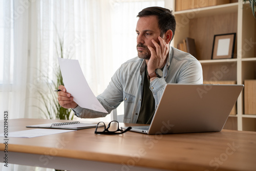 Thoughtful serious young man lost in thoughts in front of laptop, focused businessman thinking of problem solution, worried puzzled man looking at paper report