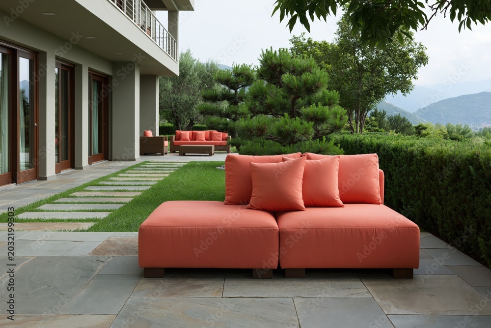 Obraz premium Modern orange modular outdoor sofa with cushions on stone patio beside manicured hedge and mountain view for terrace seating and contemporary exterior design
