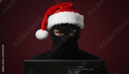 A person wearing a black balaclava and Santa hat looks at a laptop screen, symbolizing a holiday cyber threat or digital festive mischief