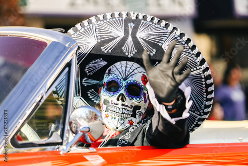 Fotografija EL PASO, TEXAS – NOVEMBER 01, 2025: A festival participant wearing a detailed sugar skull mask and embroidered mariachi sombrero waves from a classic red car during a Dia de los Muertos celebration