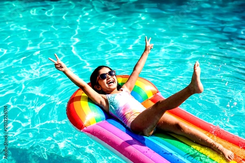 Joyful Summer Splash: A young girl in sunglasses and swimwear revels in a sun-drenched swimming pool, joyfully floating on a vibrant rainbow inflatable.