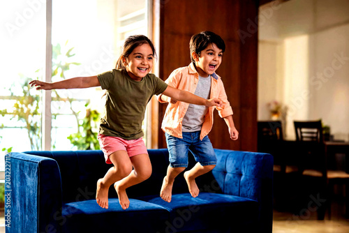Joyful Kids Jumping on Sofa: Two energetic children, a boy and a girl, leap with delight on a plush blue sofa in a brightly lit living room.