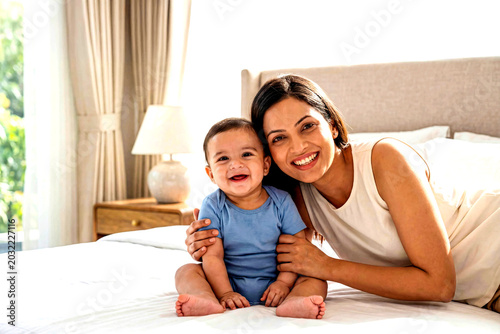 Mother and Baby Joy: A radiant mother beams as she holds her adorable baby, sharing a moment of pure happiness and love on a sunlit bed.