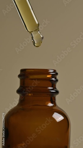 Macro shot of yellow essential oil or serum dripping from a glass dropper into an amber bottle. Natural skincare beauty product concept isolated on a neutral beige background