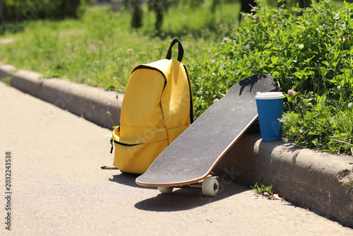 Skateboard, yellow backpack and coffee on the side of the road. Skateboard leaning on the curb next to a bright yellow backpack on a green grass background. Active lifestyle and leisure concept