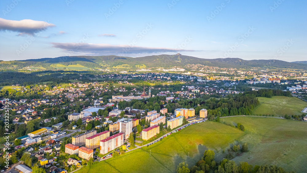 Fototapeta premium Vratislavice nad Nisou with buildings and fields. The sun shines on the town, revealing roads and greenery. Mountains are visible in the background.