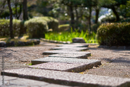 Photography A low-angle view of a stone pathway composed of rectangular granite stepping stones leading through a sunlit garden