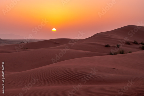 A warm sun sets over sand dunes in a desert. The landscape shows rolling dunes and a bright orange sky. Shadows stretch across the sand as night approaches.