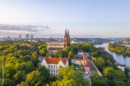 The sun rises, casting golden light over the Vysehrad Basilica of St. Peter and St. Paul in Prague. Surrounded by lush greenery, the majestic structure stands tall against the skyline.