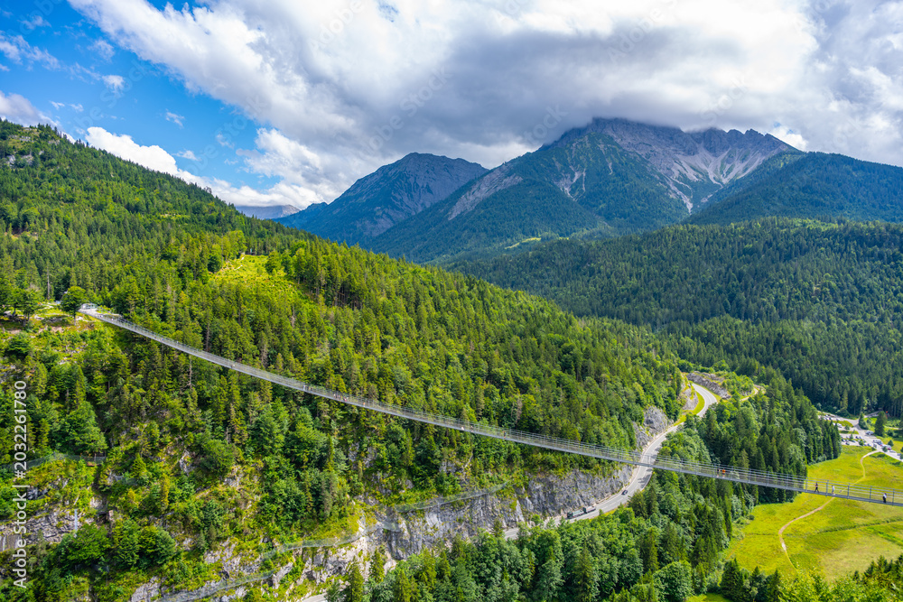 Fototapeta premium Highline 179 is a pedestrian suspension bridge in Austria. It connects a castle's ruins with a Roman fortress. Visitors walk across the bridge over a wide river gorge surrounded by mountains.