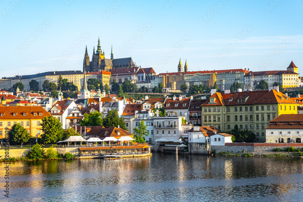Obraz premium Prague Castle stands tall above the city, with the Vltava River flowing below. Buildings line the riverbank, showing a mix of architecture against a clear sky during the day.