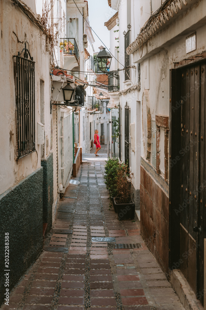 Fototapeta premium Woman in a red dress exploring a charming narrow street, showcasing traditional architecture with white walls and a brick path in, Marbella, Andalucia, Spain