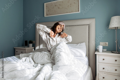 Female sitting in bed in modern bedroom with soft natural light. Concept home lifestyle, wellness and peaceful morning.
