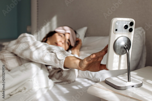 Sleepy woman stretching hand toward phone alarm while lying in bed at home. Concept waking up, morning routine and technology lifestyle.
