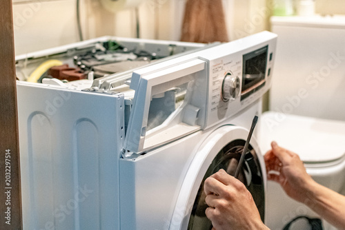 A front-loading washing machine repair in progress. The technician (or owner) is performing maintenance at home, judging by the bathroom setting in the background.