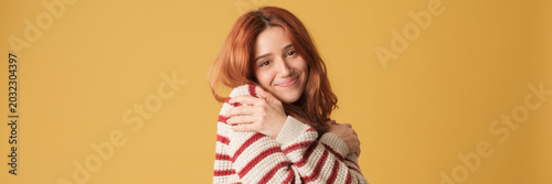 Canvastavla Cute red-haired woman dressed in sweater hugs herself looking at camera on yellow background