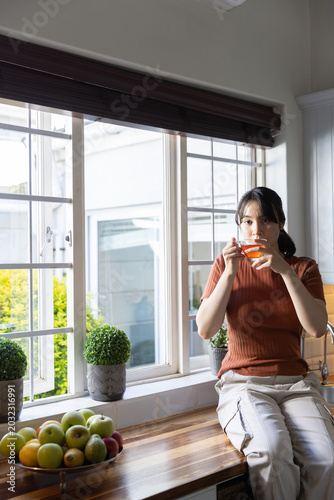 Asian woman holding teacup sipping tea on kitchen counter by window with apple bowl, copy space