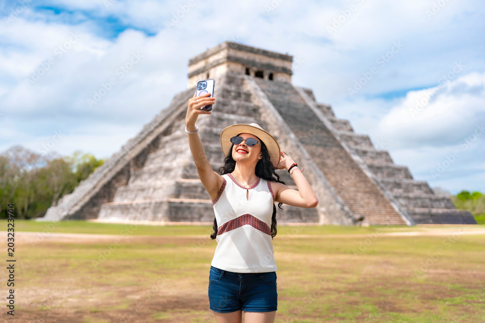 Obraz premium Young smiling Latina woman happily taking a selfie, expressing joy excitement while visiting Kukulkan pyramid at Chichen Itza, Yucatan Mexico, wearing summer outfit with hat, sunglasses in sunny day