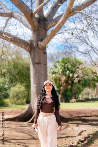 Young woman walks happily smiling relaxed near a large ceiba tree in Uxmal Yucatan Mexico, enjoying nature and cultural surroundings, wearing casual outfit with cap, sunglasses, long sleeve top pants